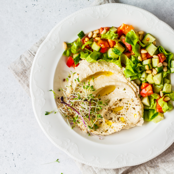 a plate of creamy hummus topped with microgreens and drizzled with olive oil, next to a side of garden salad made with romaine lettuce, tomatoes, cucumbers, onions, and white beans
