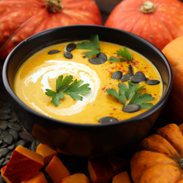 A bowl of pumpkin soup, garnished with italian parsley and pepitas, surrounded by pumpkins