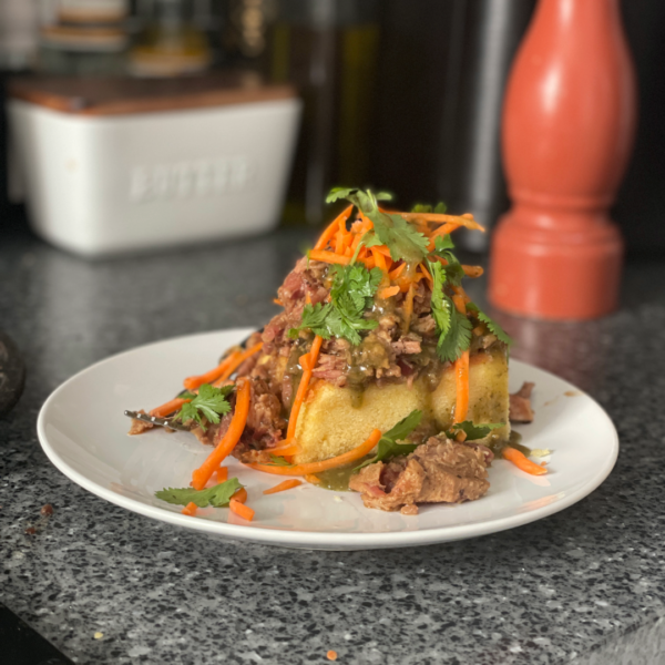 Pulled pork, shredded carrot and cilantro tops a slice of cornbread on a plate. A pepper mill and butter dish are in the background.